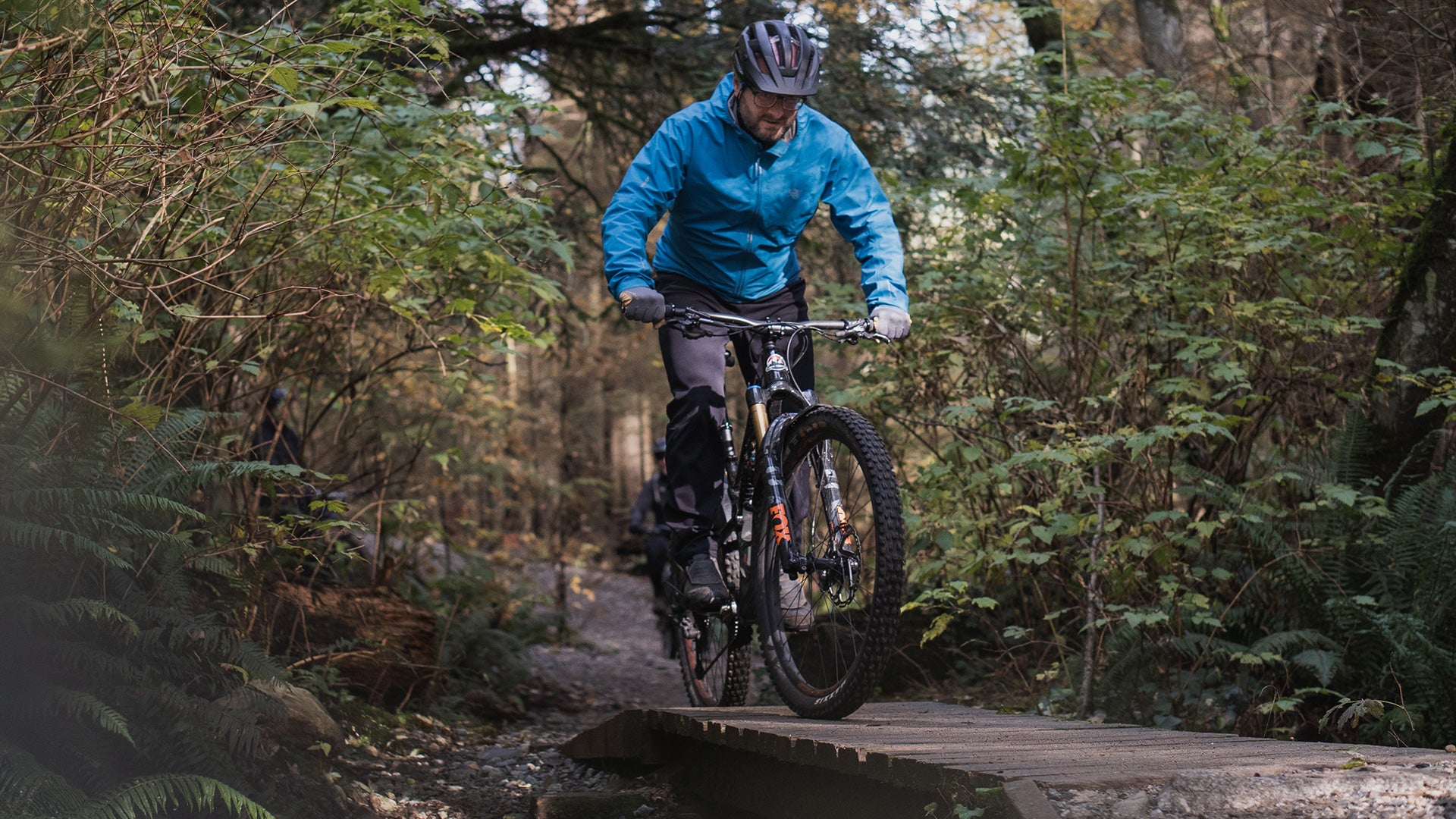 A mountain biker wearing a bright blue jacket, black pants, and a black helmet rides a full-suspension bike over a wooden bridge on a forest trail.